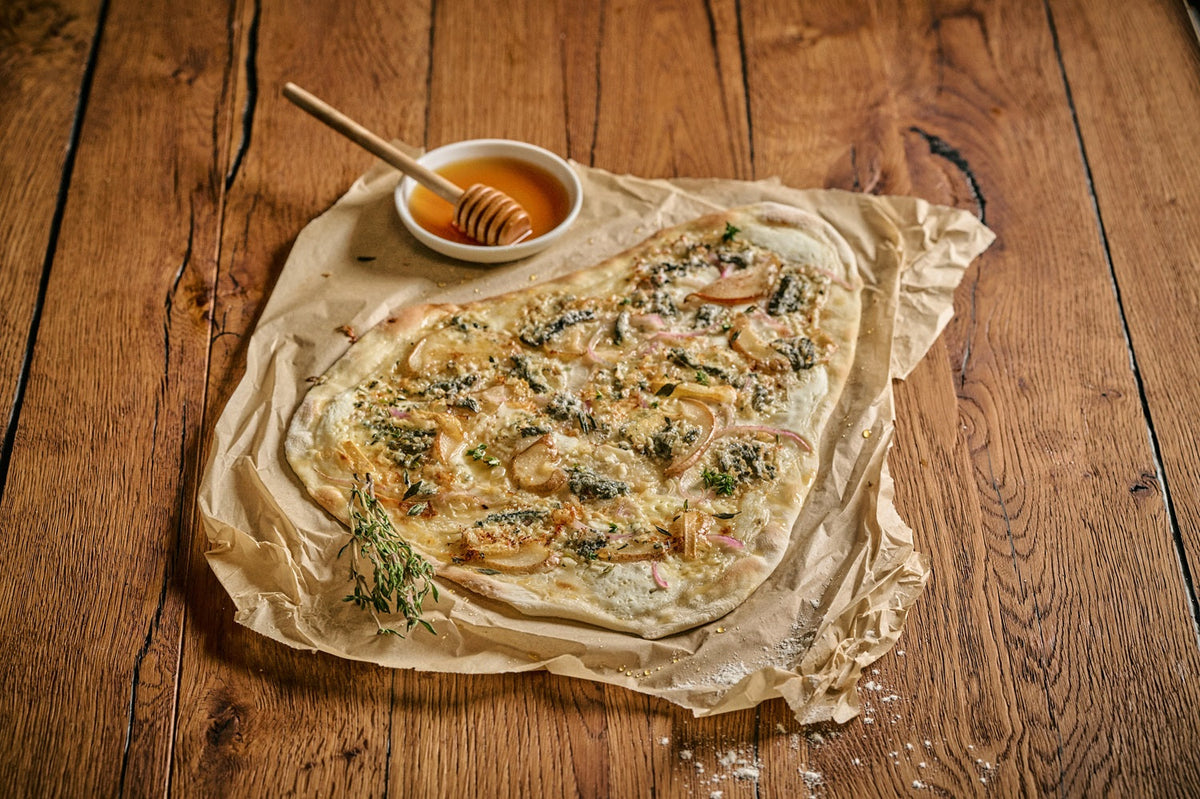 Ein Flammkuchen mit Blauschimmelkäse liegt auf einem rustikalen Holztisch, im Hintergrund liegt eine Schüssel und ein Löffel mit Honig.