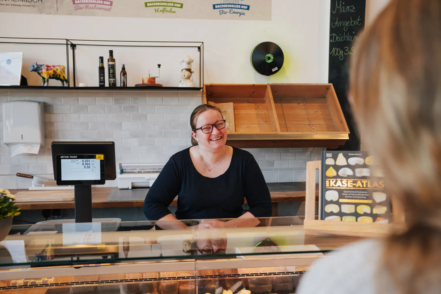 Anne steht hinterm Käsetresen und lächelt die Kundin an