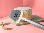 Käsehobel mit Backensholz Logo liegt vor Laib Deichkäse auf einem Schneideholzbrett.