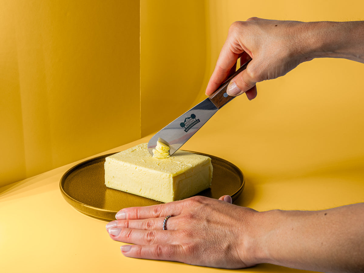 Buttermesser mit Backensholz Logo wird benutzt um Butter aufzunehmen von einem Stück, was auf einem braunem Teller liegt.