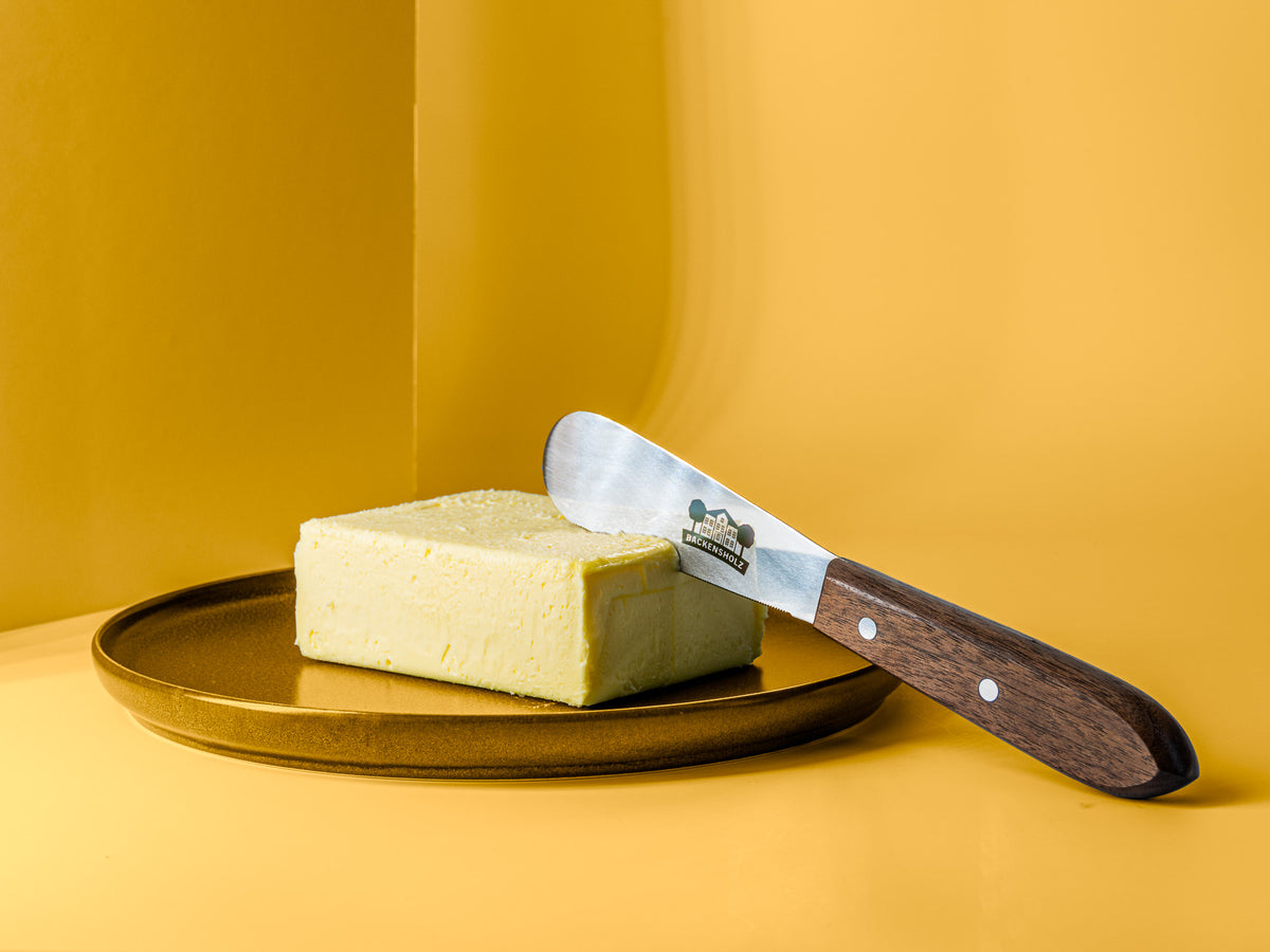 Buttermesser mit Backensholz Logo steck in Stück Butter, das auf braunem Teller liegt.