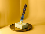 Buttermesser mit Backensholz Logo steckt in Stück Butter, das auf braunem Teller liegt.