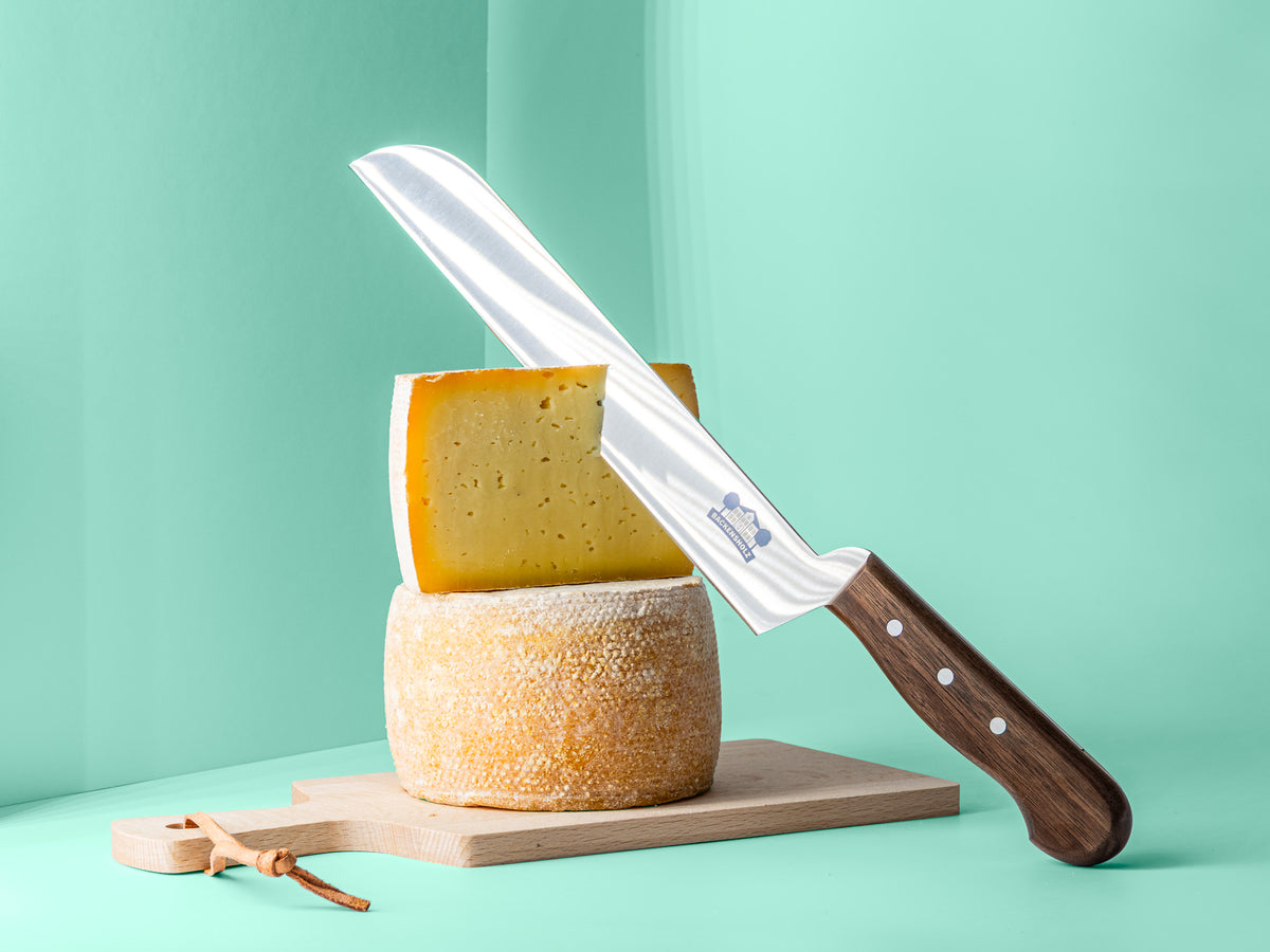 Käsemesser mit Backensholz Logo schneidet in halben Deichkäse, der auf einem ganzen Deichkäse Laib steht. Darunter liegt ein Holzbrett.