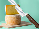 Käsemesser mit Backensholz Logo schneidet in halben Deichkäse, der auf einem ganzen Deichkäse Laib steht. Darunter liegt ein Holzbrett.