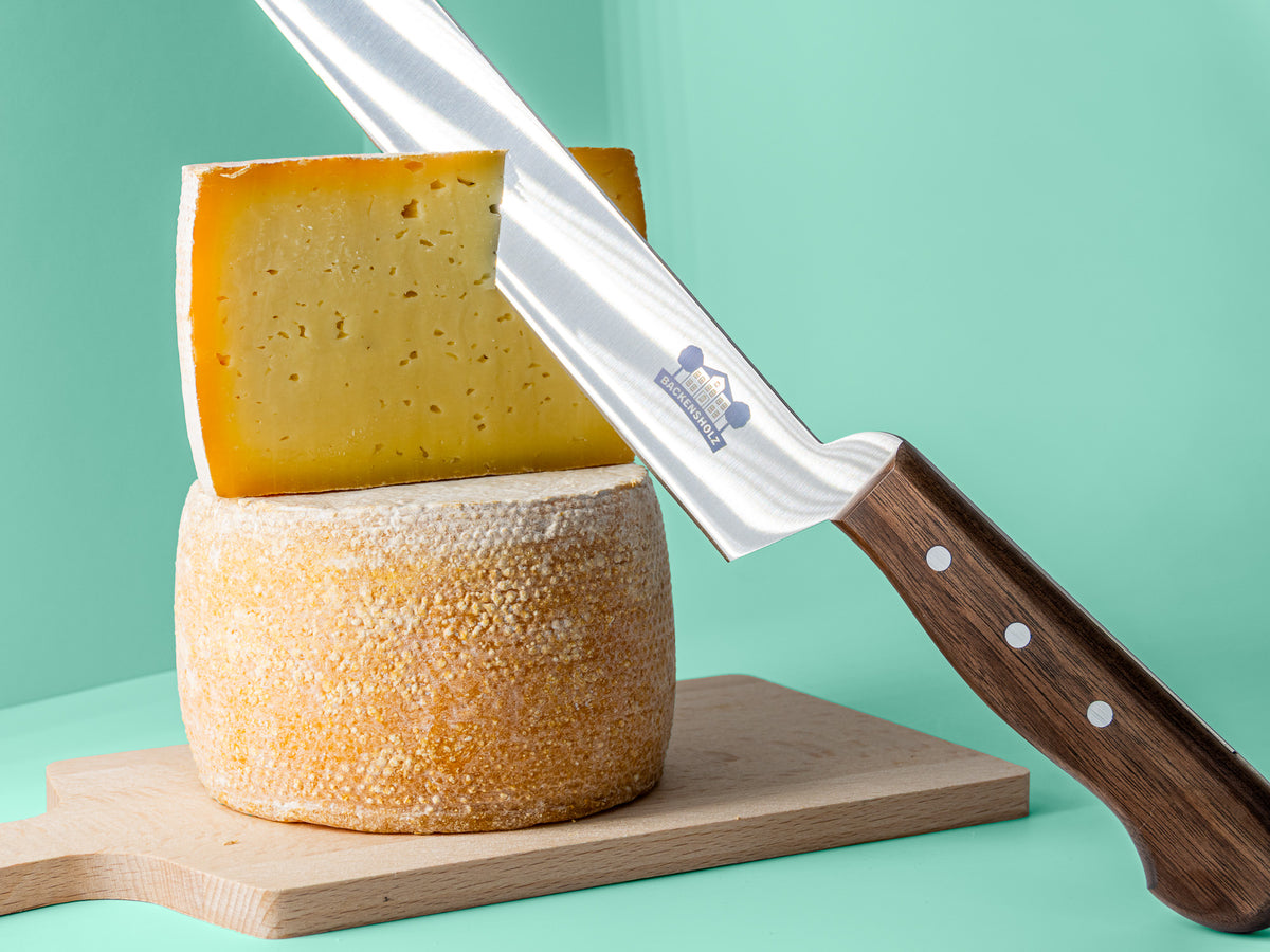 Käsemesser mit Backensholz Logo schneidet in halben Deichkäse, der auf einem ganzen Deichkäse Laib steht. Darunter liegt ein Holzbrett.